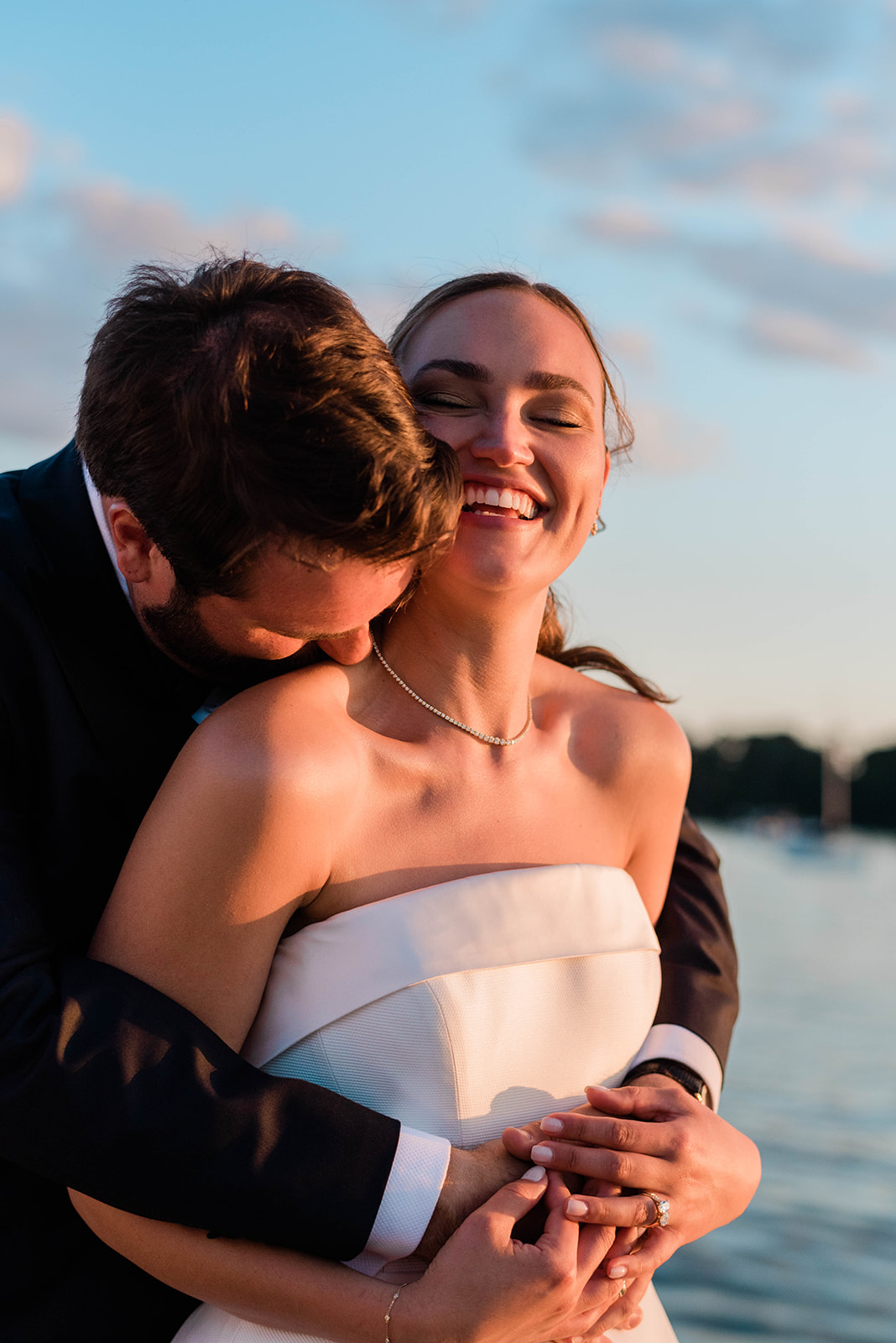groom wraps his arms around bride as they stand by the water 