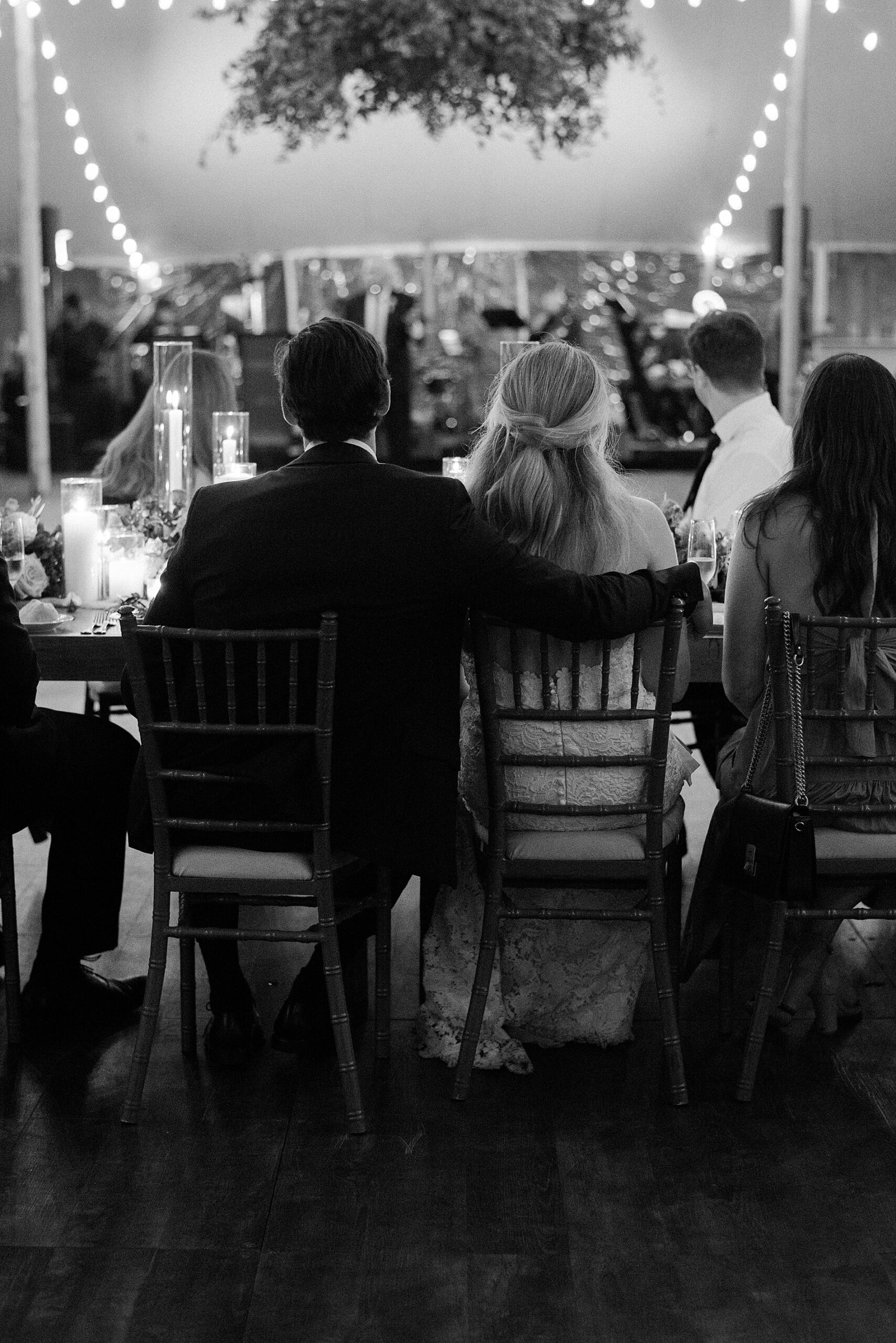 couple sit together at wedding as groom puts his arm around bride