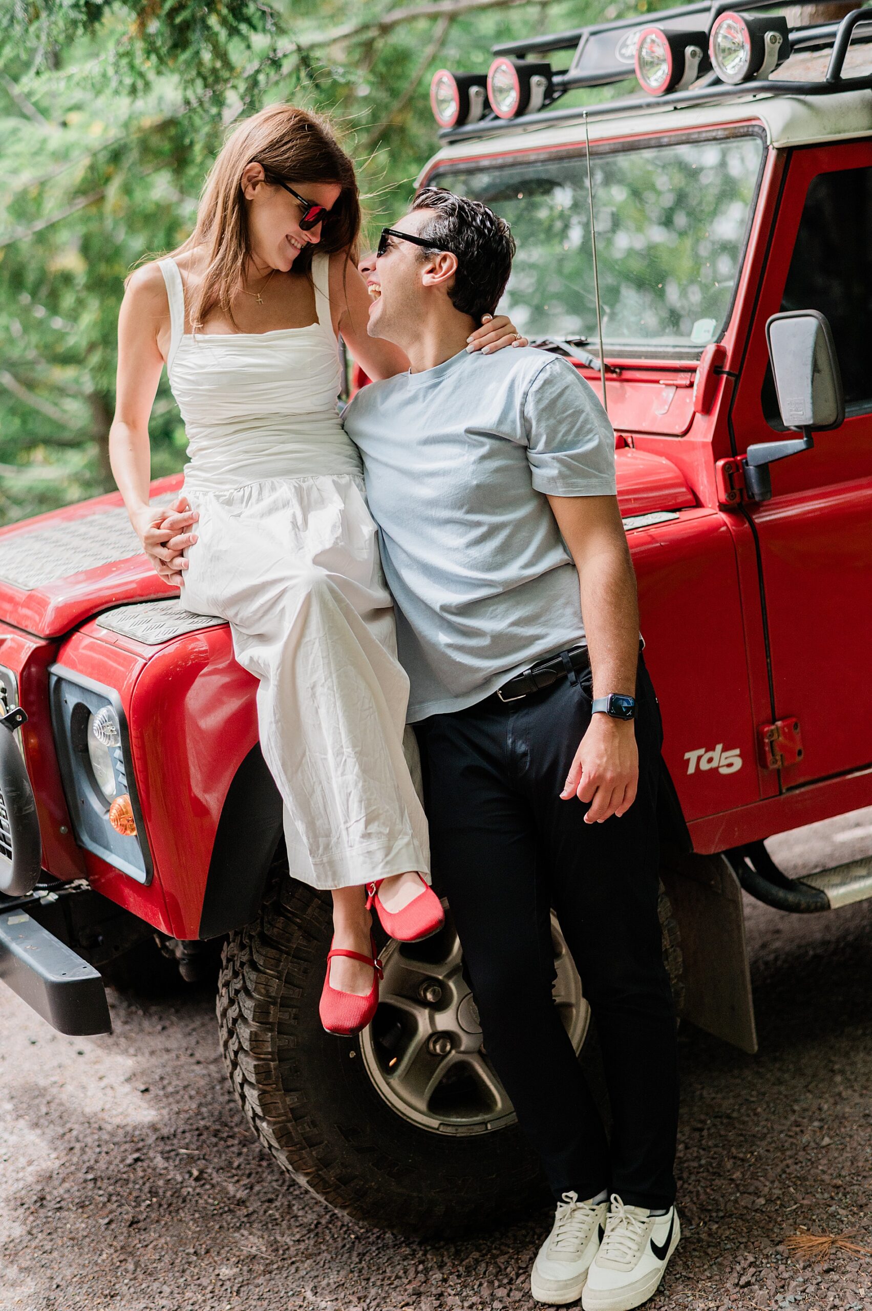 woman sits on hood of red jeep with fiance after proposal 